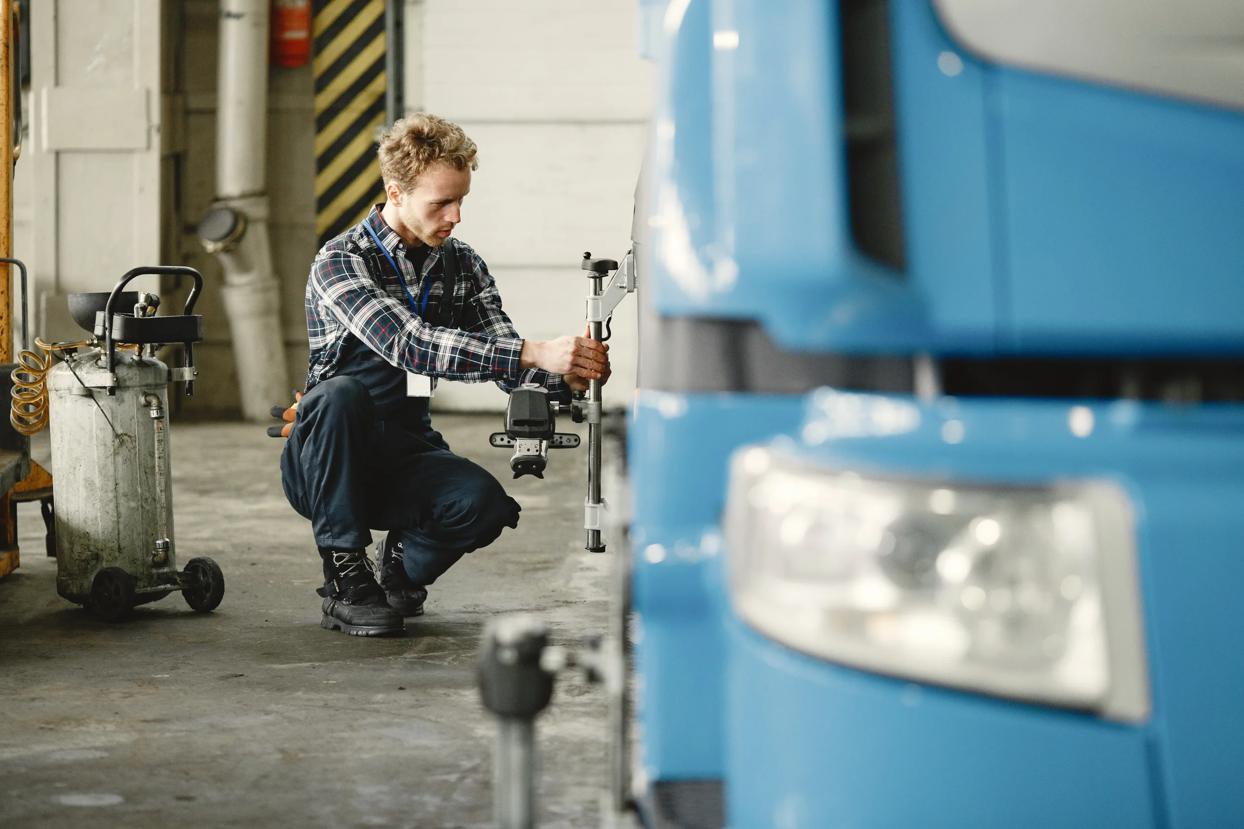 A technician in a plaid shirt kneels beside a blue truck, using a measuring tool in a garage setting.