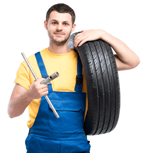 A mechanic in blue overalls holds a tire on his shoulder and a tool in his hand, smiling at the camera.