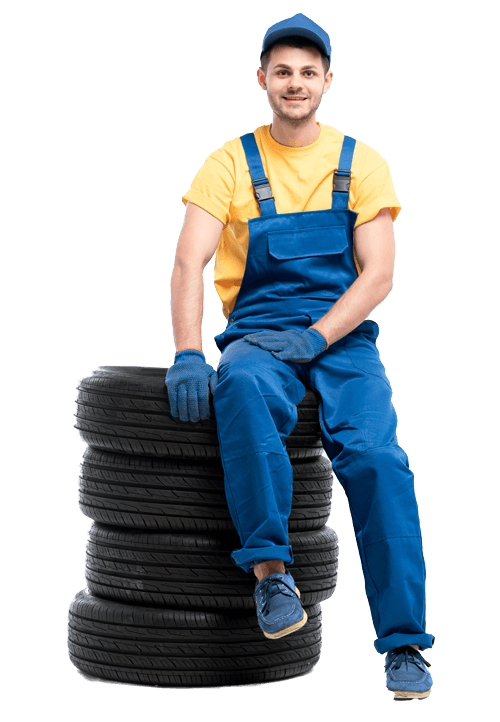 A smiling mechanic in blue overalls and gloves sitting on a stack of black tires, wearing a yellow t-shirt and a blue cap.