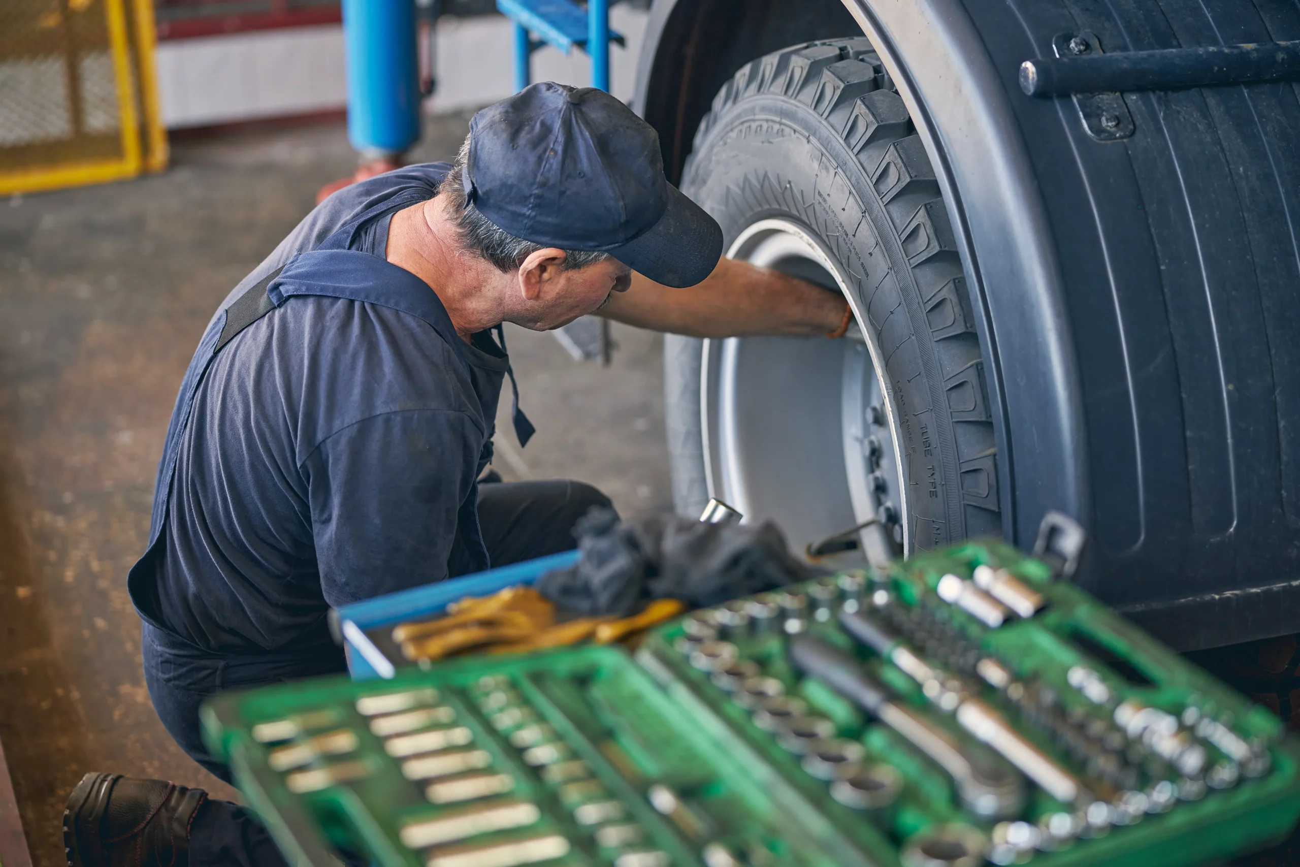 A mechanic in a cap inspects a large truck tire inside a garage, with a green tray of assorted tools visible in the foreground.
