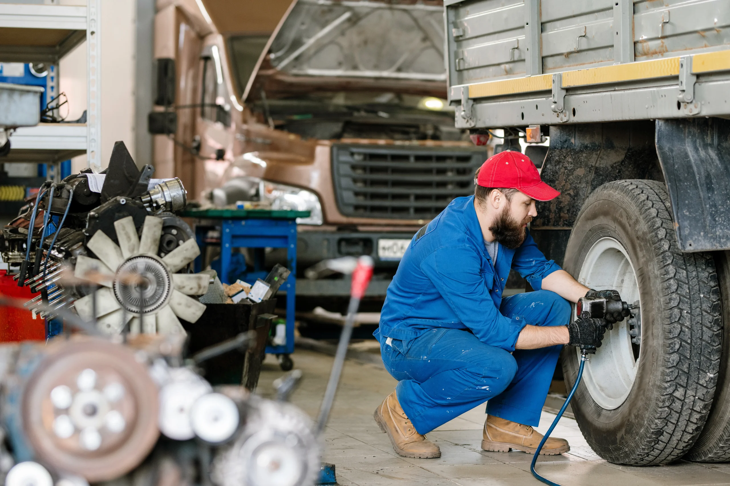 A mechanic in blue coveralls and a red cap uses a power tool to work on a truck tire in an auto repair shop. A vehicle with its hood up is visible in the background.
